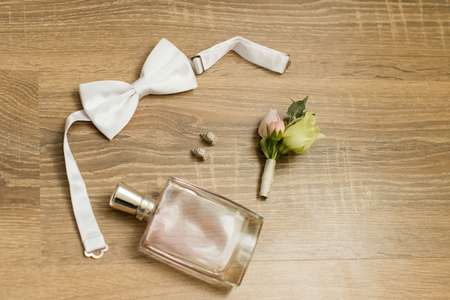 Wedding accessories. Pink perfume, bridal earrings with diamonds, white bowtie and boutonniere with small roses and a wooden backgroundの写真素材