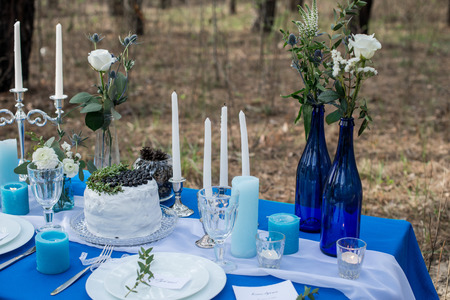 Wedding banquet table decorated with white wedding rustic cake with blueberry and greenery. Table served with cutlery, stemware and candles on a blue tableclothの写真素材