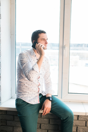 Communicative serious young businessman in a white shirt with patterns and green pants talks cell phone sitting on the windowsillの写真素材
