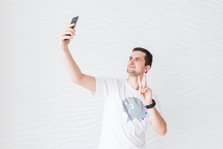 Young man in white T-shirt and blue jeans makes selfie with black smartphone on a white background and shows gesture Â«peaceÂ»の写真素材