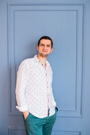 A handsome man in a white shirt with patterns and green pants leaned against the blue wall and and looking at cameraの写真素材