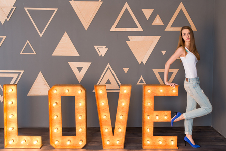 A girl in white singlet and blue denim with holes leaning on a wooden lettering Love with light bulbs on a grey backgroundの写真素材