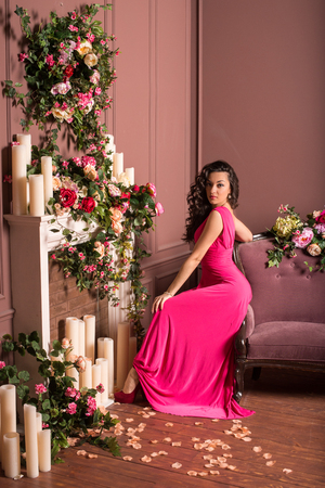 Beautiful brunette girl with long curly hair wearing a pink dress. Girl sitting on the violet sofa. A lot of flowers and candles in the room.の写真素材