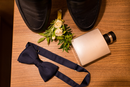 Perfume in a metal bottle, blue bow tie and boutonniere with small roses and a part of black shoes on a wooden backgroundの写真素材