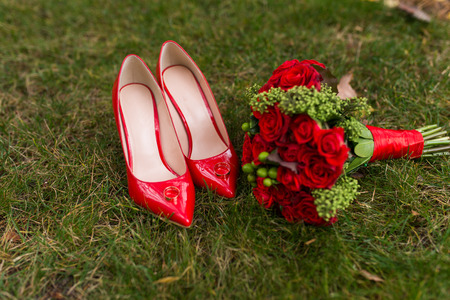 Women's fashion red wedding shoes with golden rings on them in focus and bride's bouquet with red roses and green berries.の写真素材