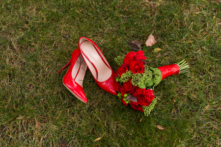 Female fashion red wedding shoes with bride's bouquet of red roses and green berries on green grass background.の写真素材