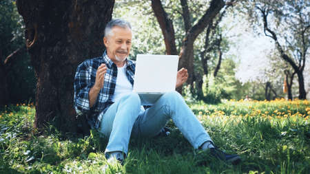 An elderly gray-haired man with a laptop is excited and rejoices at an online victory or good news, sitting on the grass among flowers in a city park on a sunny summer day.の写真素材