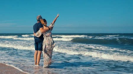 A happy slender romantic middle-aged couple enjoying a relaxing walk on a sandy beach on a sunny day, against the background of large sea waves.の写真素材