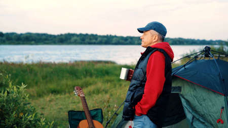 An elderly male tourist with an acoustic instrument enjoys nature and the sunset sunlight. Lifestyle, the concept of rest in old age. An elderly slender man with a guitar by the river at sunset.の写真素材