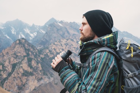 A hipster man with a beard in a hat, a jacket, and a backpack in the mountains holds binoculars, adventure, tourism, trackingの写真素材