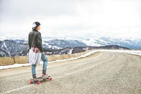 Stylish red-haired girl in a cap and denim overalls standing on a bump at the highway in the mountains with a longboardの写真素材