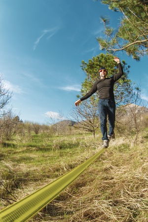 A man, aged with a beard and wearing sunglasses, balances on a slackline in the open air between two...
