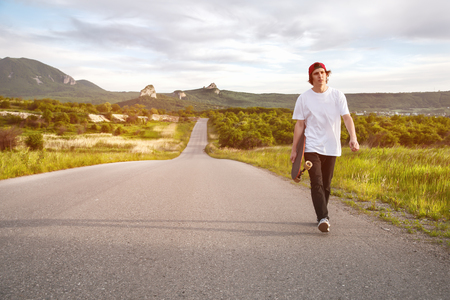 A man in a cap and with a long board in his hands is walking on a country roadの写真素材