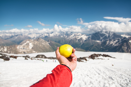 A mans hand holds an apple against the background of snow-capped mountains and snow underfoot.の写真素材