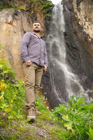 A bearded hipster in a plaid shirt stands against a waterfall backgroundの写真素材