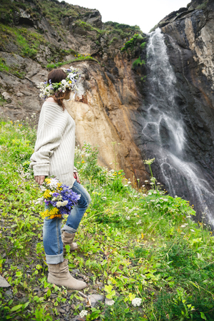 Portrait of a girl with a wreath on her head and a bouquet of flowers in her hands against a waterfallの写真素材