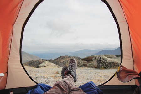 Legs of a man resting in boots for mountain tracking in travel tent against the backdrop of mountains and valleys with noisy cloudsの写真素材