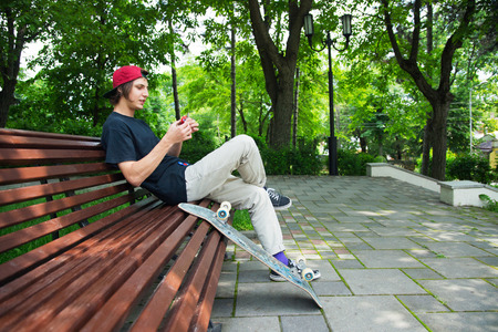 Long-haired hipster skater in a cap sits on a bench and spins on a spin-spinnerの写真素材