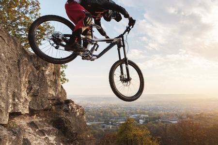 a young rider at the wheel of his mountain bike makes a trick in jumping on the springboard of the downhill mountain path in the autumn forestの写真素材