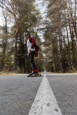 A young hipster in a cap and plaid shirt is riding his longboard on a country road in the forestの写真素材