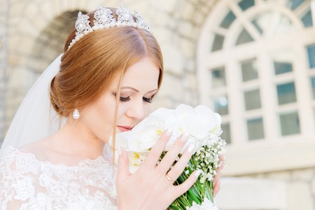A young bride in a white dress is sniffing her wedding bouquet that is carefully held by her handsの写真素材