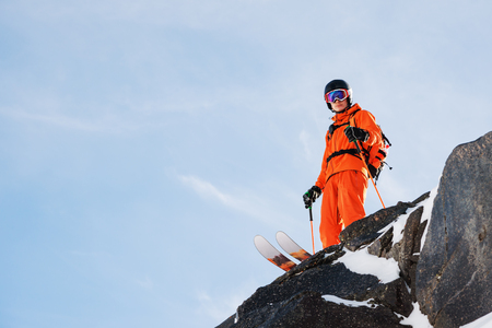 Professional skier-athlete stands on the edge of a high cliff against a blue sky on a sunny dayの写真素材