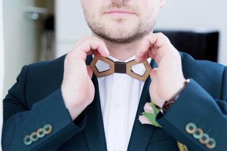 A sturdy newlywed with a beard in a suit adjusts the wooden bow tie on the collar with his white shirt.の写真素材