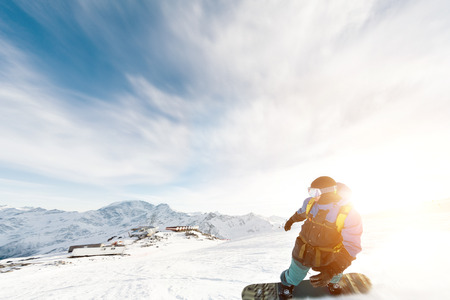 A snowboarder in a ski mask and a backpack is riding on a snow-covered slope leaving behind a snow powder against the blue sky and the setting sun