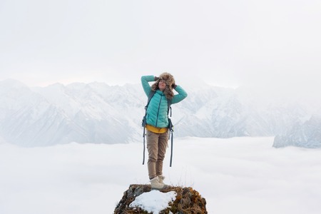 A happy smiling hipster girl travels in a down jacket with a backpack and in a large fur hat stands on top of a rockの写真素材