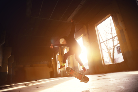 A young skater in a white hat and a black sweatshirt does a trick with a skate jump in an abandoned building in the backlight of the setting sun.の写真素材