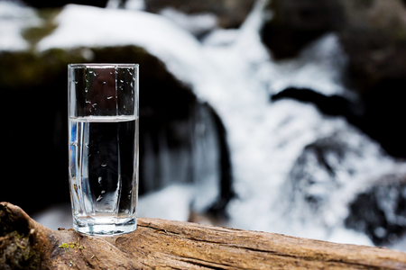 A transparent glass glass with drinking mountain mineral water stands on the wood against the backdrop of a rapid flow of a clean mountain river in winter.の写真素材