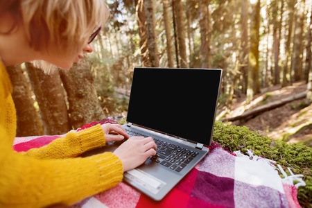 A mock-up image of a womans hand using and typing on a laptop with a blank black desktop on a plaid grass lying in the forestの写真素材