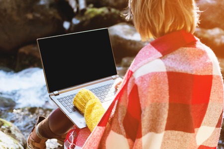 Mockup image of a girl wrapped in a plaid plaid with a laptop with a blank black desktop on her lap against the background of a mountain river and large stones.の写真素材