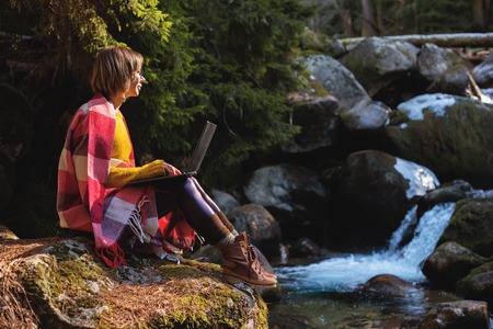 A toned portrait of a a smiling freelancer hipster girl with glasses dressed in a blanket with a laptop kneeling sitting on a rock in a coniferous forest next to a stormy mountain river in warm sunlight. The concept of freelancing work at a distance and traveling without interrupting workの写真素材