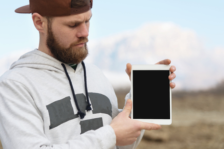 Close-up Portrait of a hipster in a brown cap in the open air holds a white tablet pc in his hands. A bearded man looks at the tablet. background blurの写真素材
