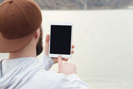 Close-up of a horde in a brown cap in the open air holds a white tablet pc in his hands.