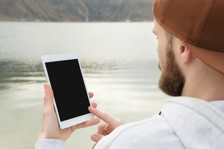 Close-up of a horde in a brown cap in the open air holds a white tablet pc in his hands. A bearded man looks at the tablet. OTS view from behind the shoulder. Makes a swap move with his fingerの写真素材