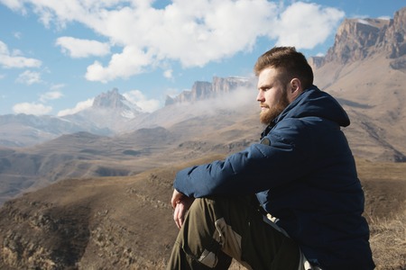 A portrait of a stylish bearded hipster sitting on a rock against the backdrop of epic rocks and contemplating into the distance thinking about life. The concept of tranquility and solitude with natureの写真素材