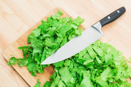 Close-up on a wooden table is a wooden cutting board on which lies the chopped lettuce leaves and on them lies a large cutting knife. The concept of self-cooking vegetarian food at home. Healthy lifestyleの写真素材