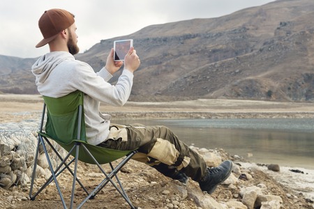 A bearded hipster in a cap sits on a folding chair on the shore of a high mountain lake and takes a...