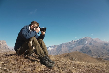 Portrait of a bearded hipster, a photographer takes pictures of his DSLR against the backdrop of snow-capped Caucasian mountains on a sunny day. The concept of a free life with tourism and photography on a journeyの写真素材