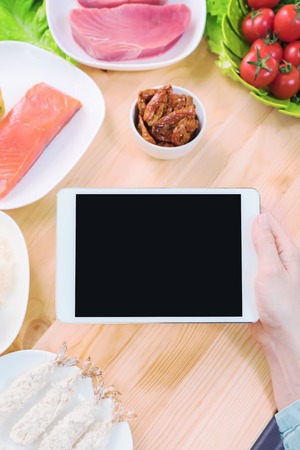 Close-up top view of female hands holding a tablet computer with a blank display surrounded by healthy food on a home kitchen table.の写真素材