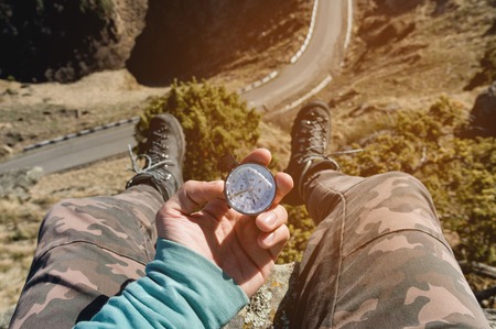 Hiker sitting on a high rock holds a compass in front of her feet in trekking boots and a high cliff...