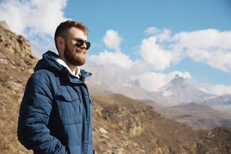 Hipster traveler in a down jacket and sunglasses stands on a mountain slope against the backdrop of epic rocks and smiles. The concept of finding happiness in the mountainsの写真素材