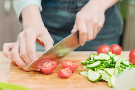 Close-up Delicate female hands cut a large knife with tomatoes on a quarter on a wooden board at home. Home kitchen. Healthy eating. The concept of vegetarianismの写真素材