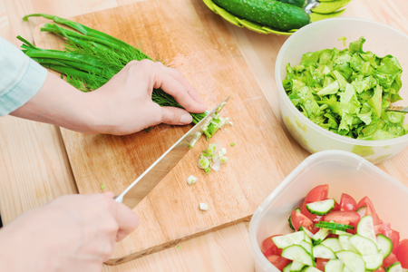 Close-up of gentle female hands slice on the wooden board the stalks of green onions making salad. The concept of a vegetarian healthy food and lifestyle. Making healthy food at homeの写真素材