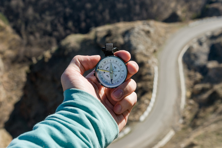 Close-up A man's hand holds a pocket compass against the backdrop of a mountain road and forest. The concept of outdoor navigation.の写真素材