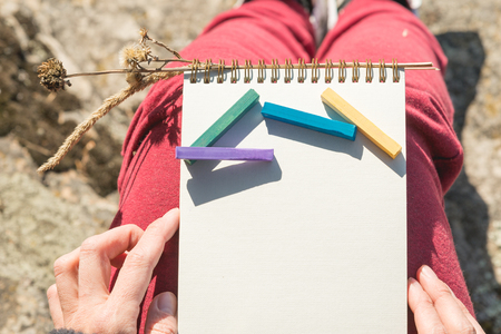 View from the first person on the female lap is a notebook with chalks for drawing pastels. Female hands holding a notebook in natureの写真素材