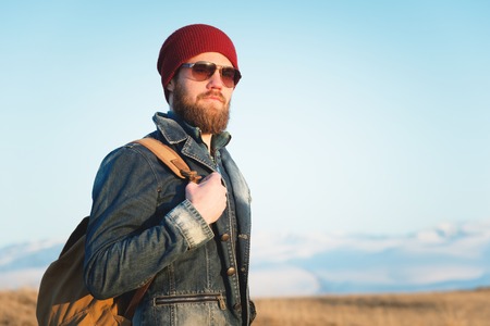 Portrait of a thoughtful stylish hipster with a beard in sunglasses and a hat with a backpack on the background of the snow-capped Mount Elbrus in the Caucasus at sunsetの写真素材