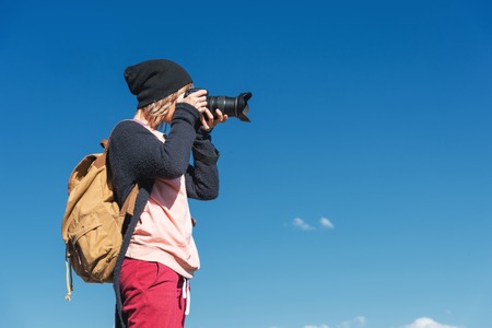 Portrait of a stylish girl hippy in a hat and with a backpack that takes pictures of her on a DSLR camera outdoors against a blue sky on a sunny afternoon. The concept of photography in travelの写真素材
